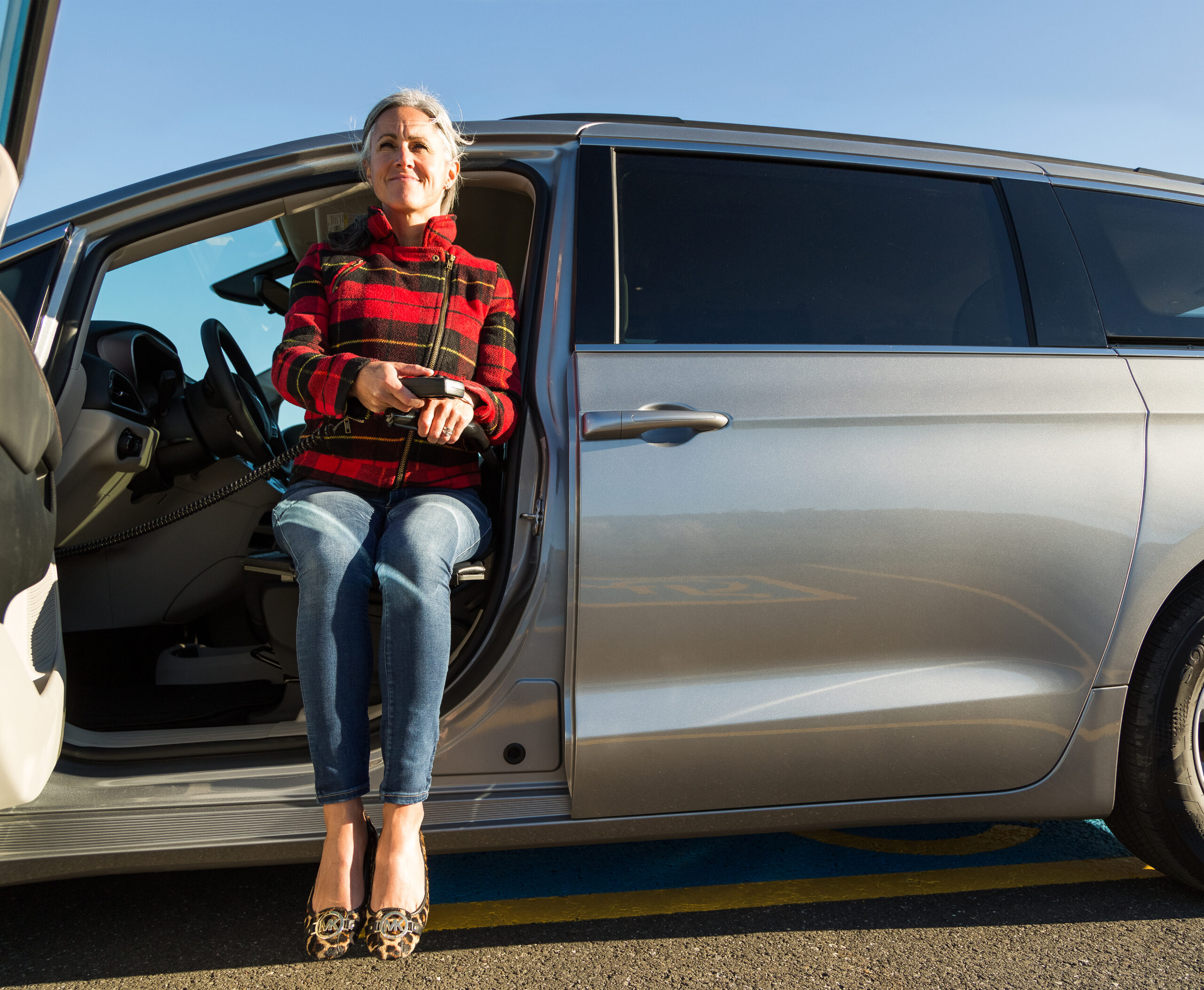Woman sitting on the open door of a silver car holding a travel mug.
