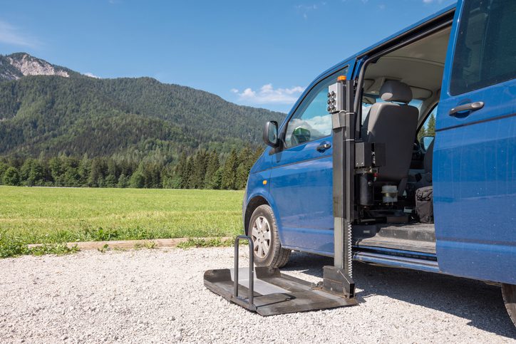 Blue van parked with wheelchair ramp extended on gravel road near fields and hills.