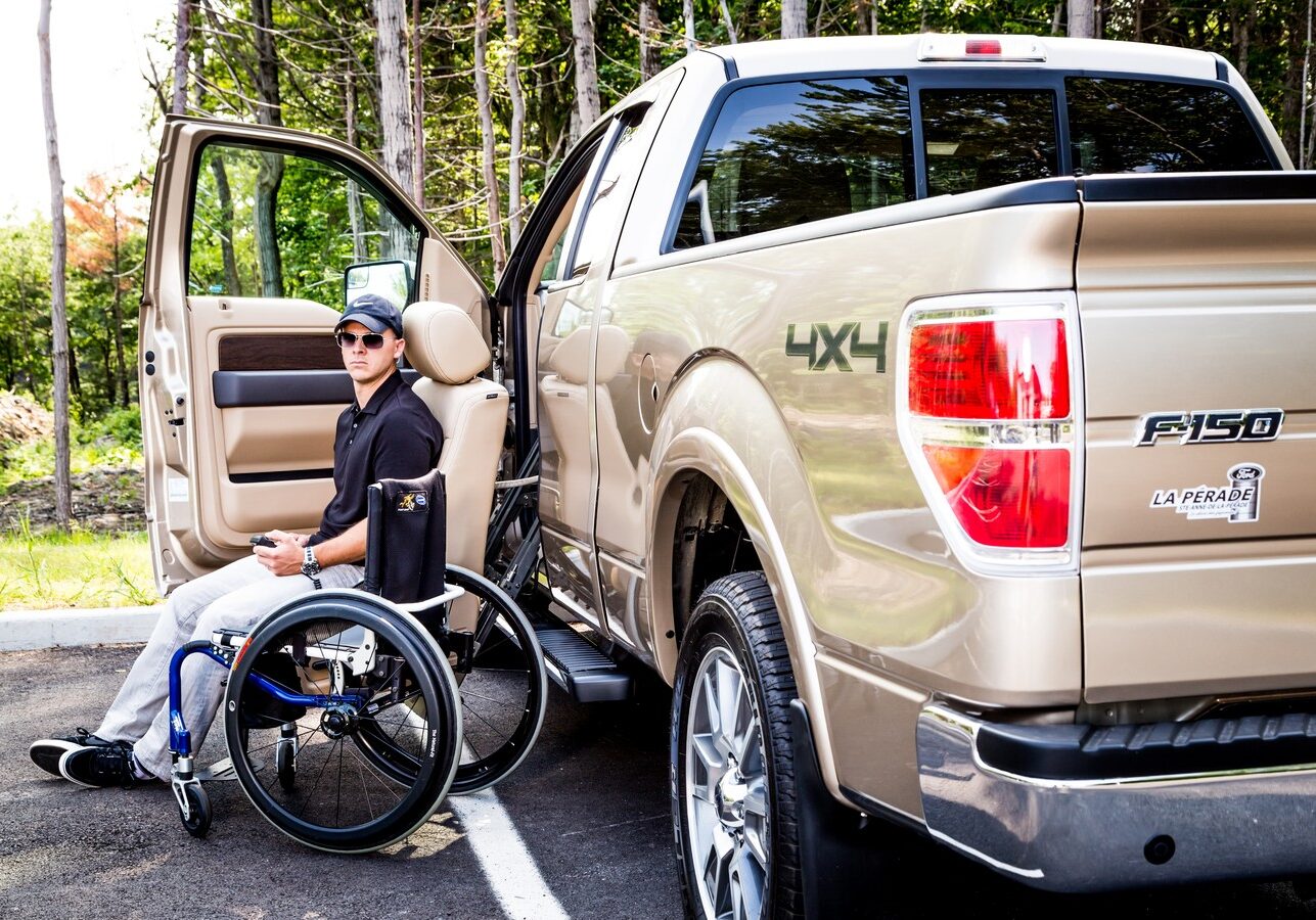 A woman in a wheelchair beside a pickup truck outdoors.